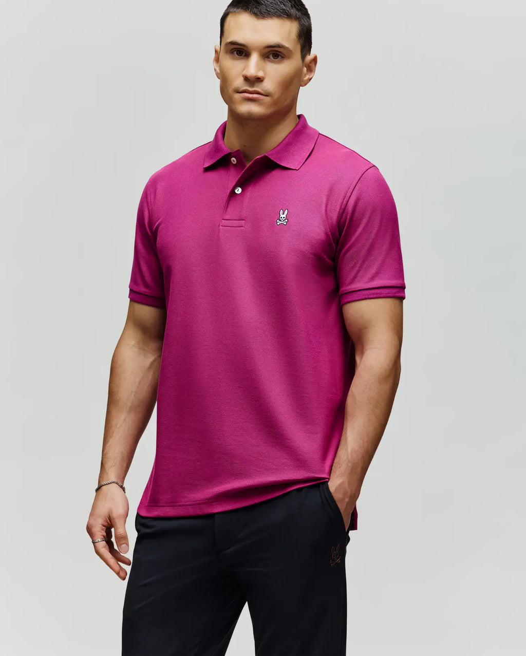 A man stands against a plain background wearing a Psycho Bunny MENS CLASSIC PIQUE POLO SHIRT - B6K001F200 in bright pink, made from soft Pima cotton with an embroidered Bunny logo on the chest, paired with black pants.