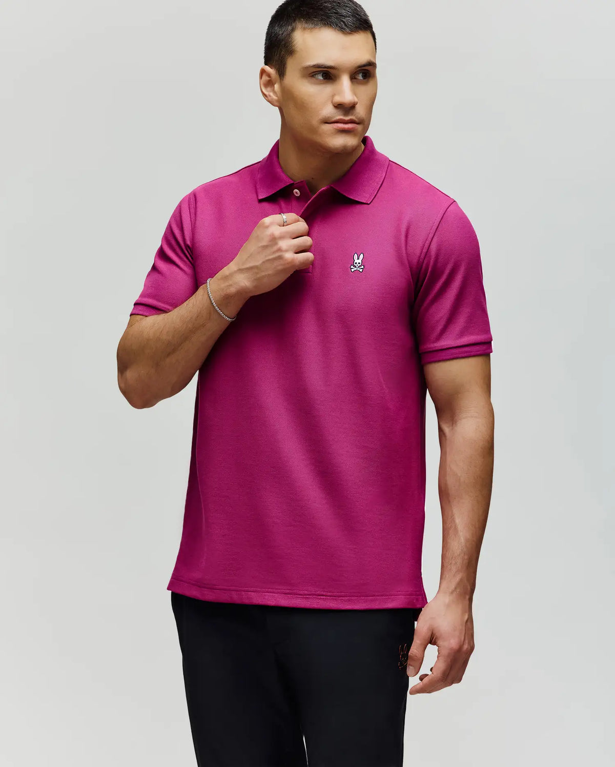 A man in a Psycho Bunny MENS CLASSIC PIQUE POLO SHIRT (B6K001F200) with an embroidered Bunny logo stands against a light background, looking left and holding his shirt collar. He has short dark hair.