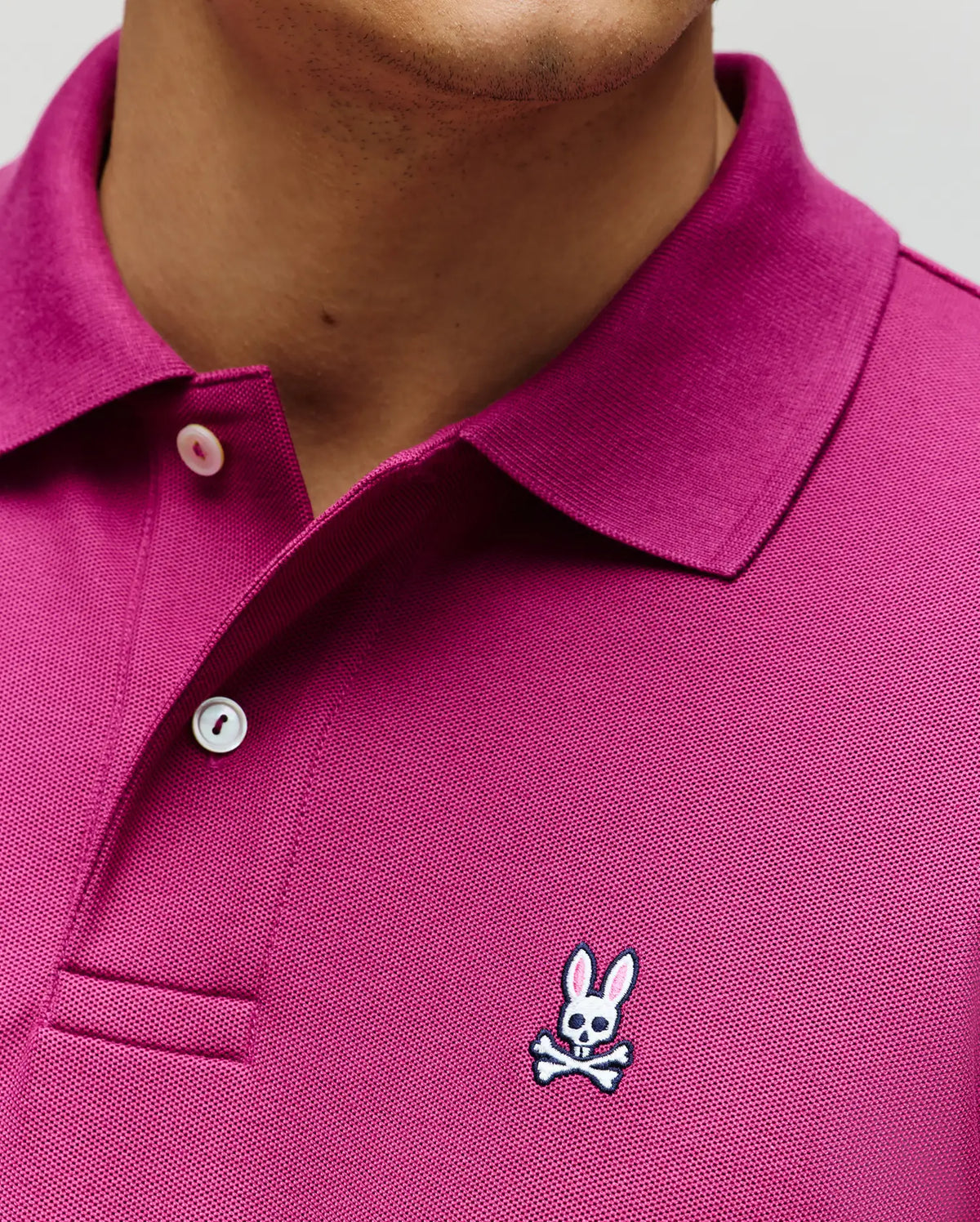 Close-up of the Psycho Bunny MENS CLASSIC PIQUE POLO SHIRT (B6K001F200) in magenta Pima cotton with white buttons and an embroidered Bunny logo—a bunny head with crossbones—on the left chest.