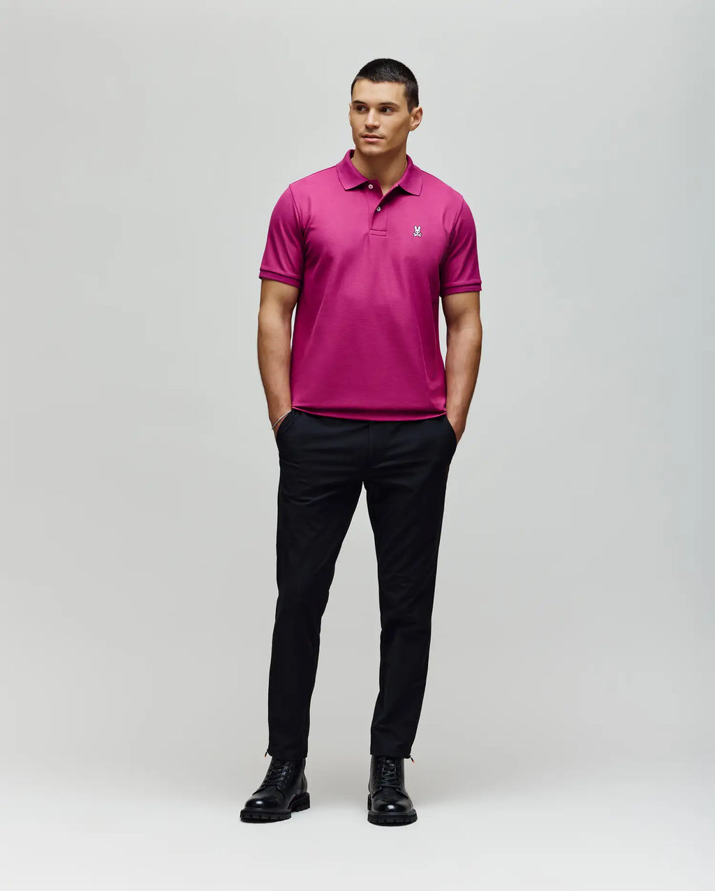 A man stands against a plain background wearing the Psycho Bunny MENS CLASSIC PIQUE POLO SHIRT - B6K001F200 in bright pink, paired with black pants and boots. His hands are in his pockets as he looks slightly to the side.