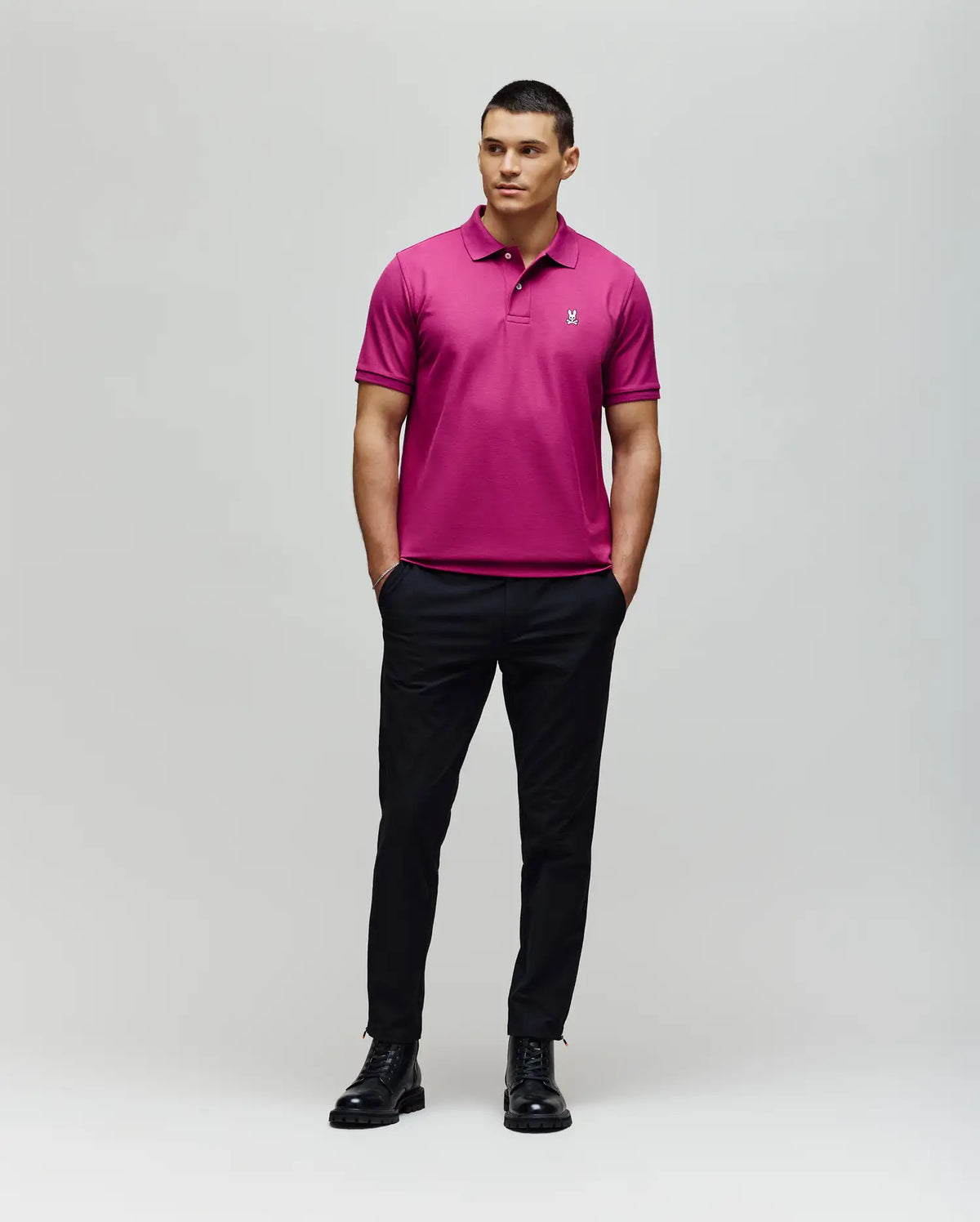 A man stands against a plain background wearing the Psycho Bunny MENS CLASSIC PIQUE POLO SHIRT - B6K001F200 in bright pink, paired with black pants and boots. His hands are in his pockets as he looks slightly to the side.