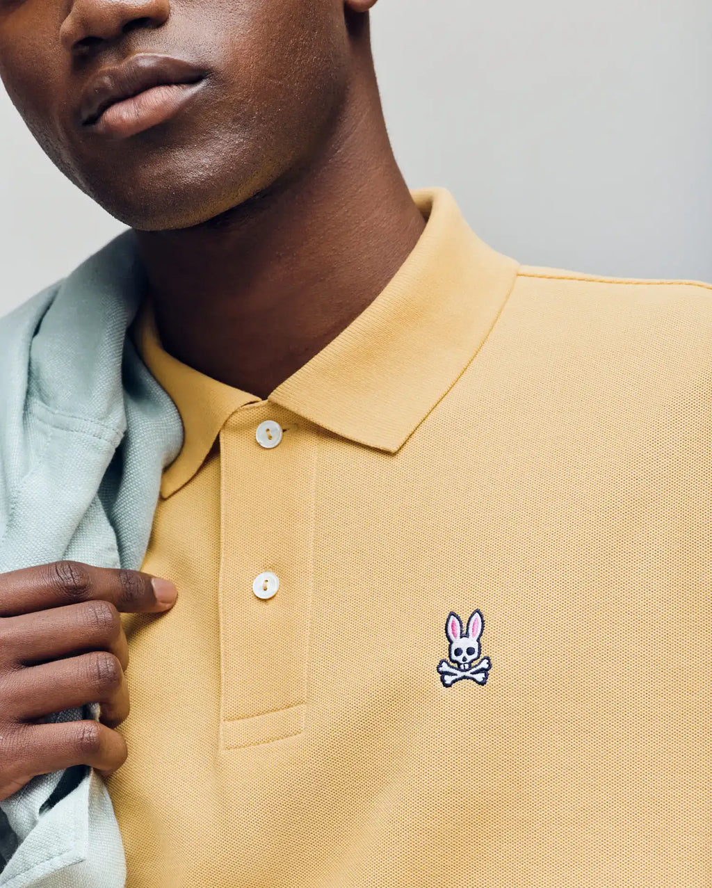 A person wears a light yellow Psycho Bunny MENS CLASSIC PIQUE POLO (B6K001F200) featuring an embroidered pink Bunny logo above white crossed bones on the chest, and holds a light garment over their shoulder.