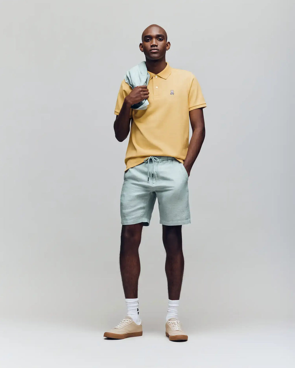 A man stands against a plain background wearing a Psycho Bunny MENS CLASSIC PIQUE POLO - B6K001F200 in yellow, light green drawstring shorts with a matching jacket over his shoulder, white socks, and beige sneakers, one hand in his pocket.