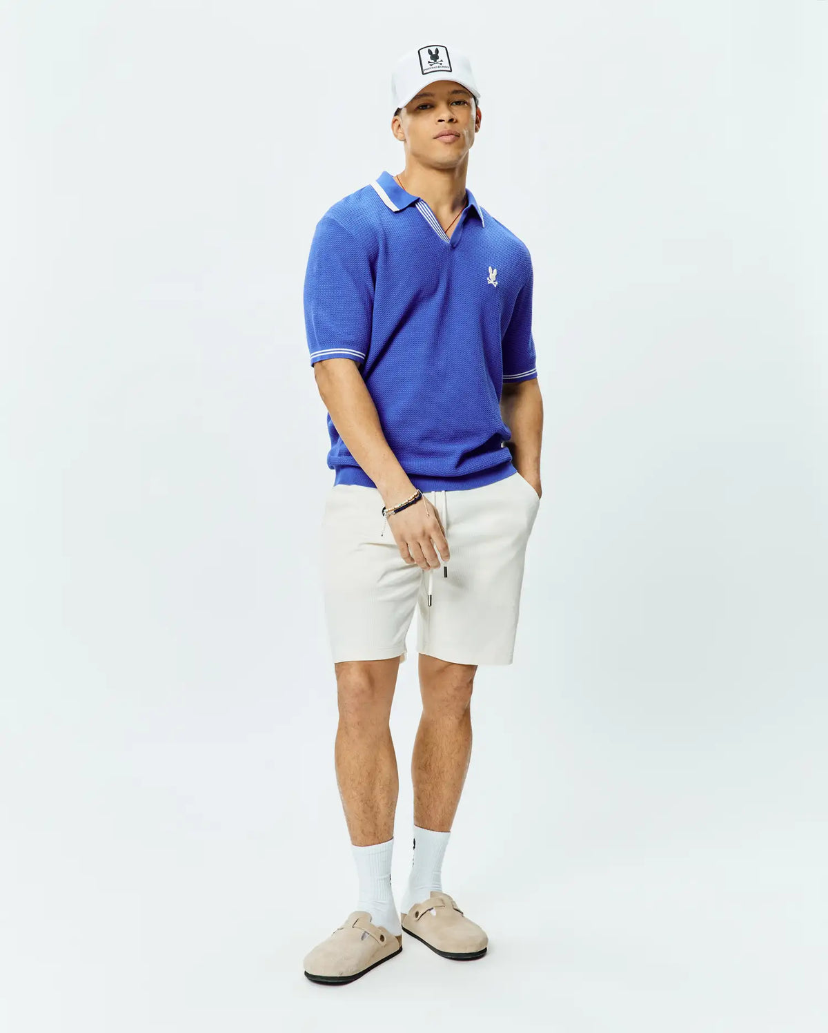 Mens Innis Polo, a vibrant blue classic fit textured knit polo with white tipped collar and cuffs, featuring a small embroidered Psycho Bunny skull logo on the left chest, and relaxed off-white textured drawstring shorts.