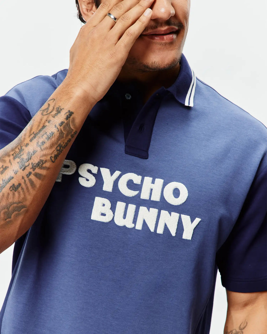 Mens Kelly Oversized Polo, a color-block jersey polo in faded blue with navy sleeves and collar, featuring white collar stripes and a large textured white appliqué PSYCHO BUNNY logo on the chest.