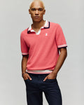 A man with short blond hair wears the Psycho Bunny MENS CARTER SWEATER POLO - MT0400093 in pink, featuring a Johnny collar and white/navy trim, paired with navy pants and a bracelet, standing against a plain light background.