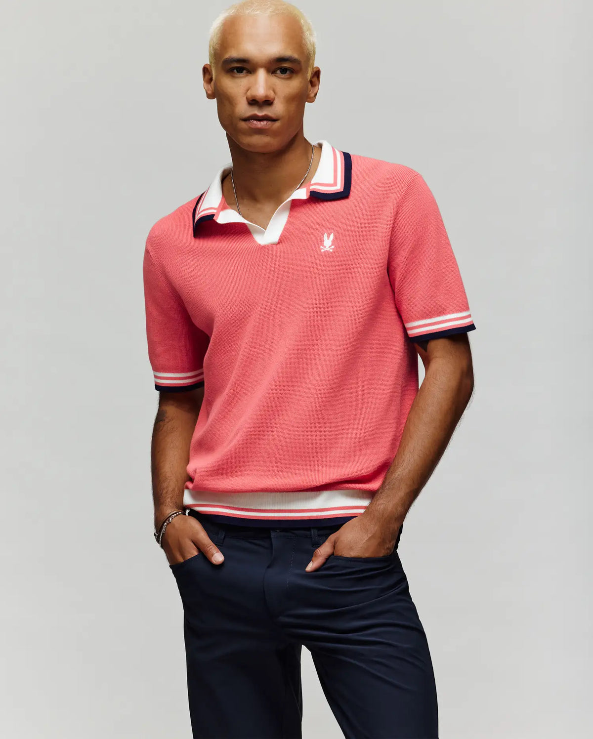 A man with short blond hair wears the Psycho Bunny MENS CARTER SWEATER POLO - MT0400093 in pink, featuring a Johnny collar and white/navy trim, paired with navy pants and a bracelet, standing against a plain light background.
