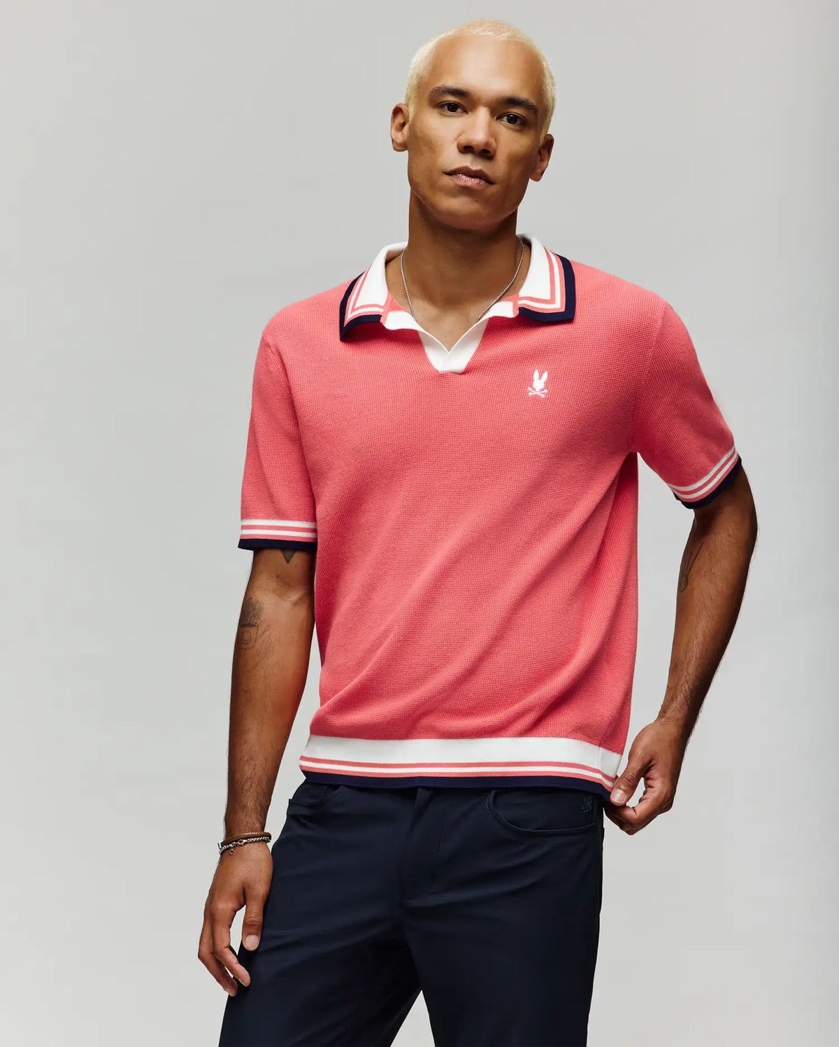 A man with short bleached blond hair wears the Psycho Bunny MENS CARTER SWEATER POLO (MT0400093), a coral pink, regular fit cotton knit featuring a Johnny collar and white/navy striped trim, paired with dark pants against a light gray background.