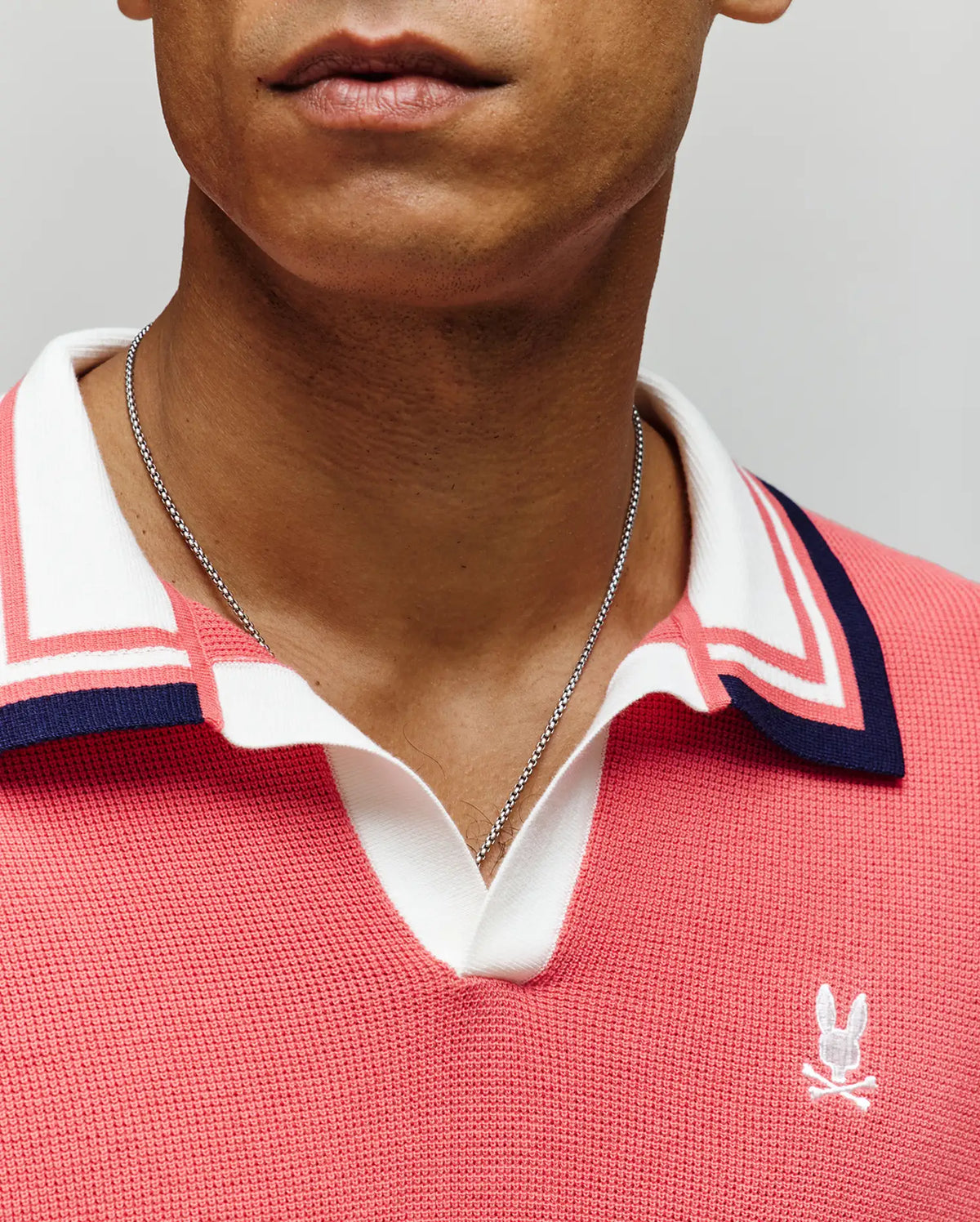 A person wearing a silver chain necklace and the Psycho Bunny MENS CARTER SWEATER POLO (MT0400093) in coral pink with white and navy Johnny collar details and a white embroidered bunny logo. Image shows lower face and upper torso only.