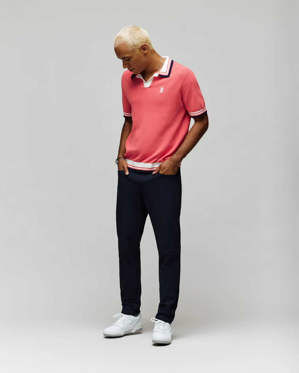 A person with short, bleached blonde hair models the Psycho Bunny MENS CARTER SWEATER POLO (MT0400093) in pink, paired with dark pants and white sneakers, standing against a light gray background and looking down with hands in pockets.